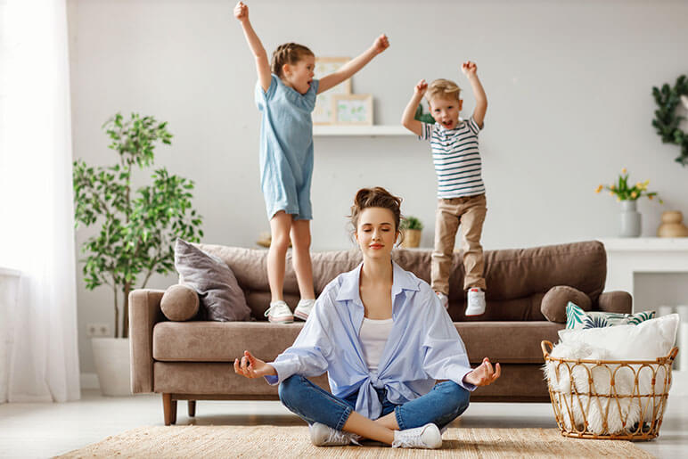 A mom meditated on the ground while her two kids are jumping on the couch behind her