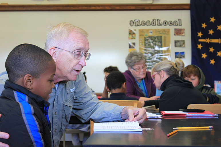 Older man helping child read at a school desk in a school