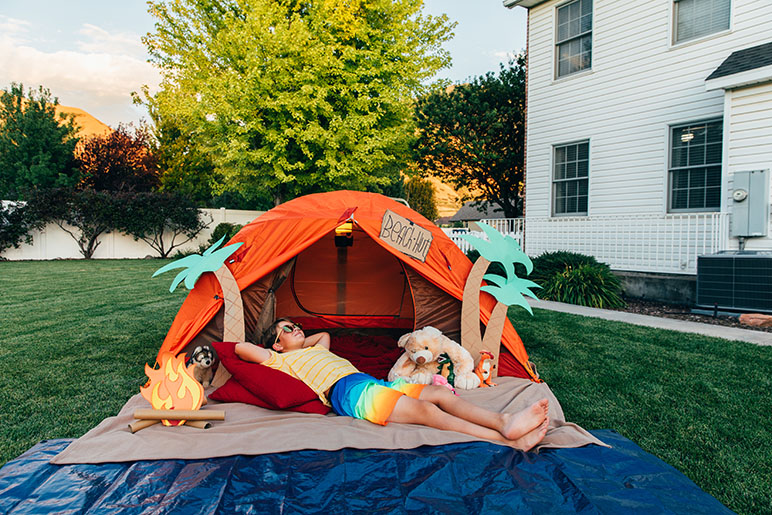 Young boy laying down on a blanket in a tent outside with fake palm trees and stuffed animals around him